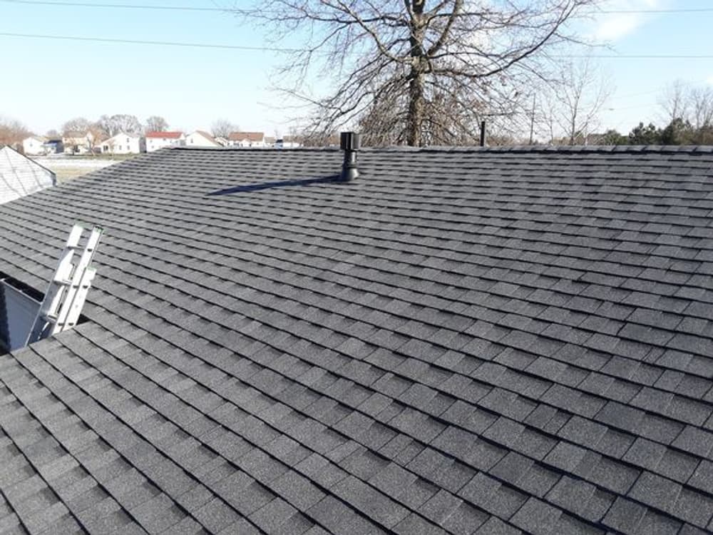 Asphalt shingle roof with chimney and ladder against a clear sky and residential background.