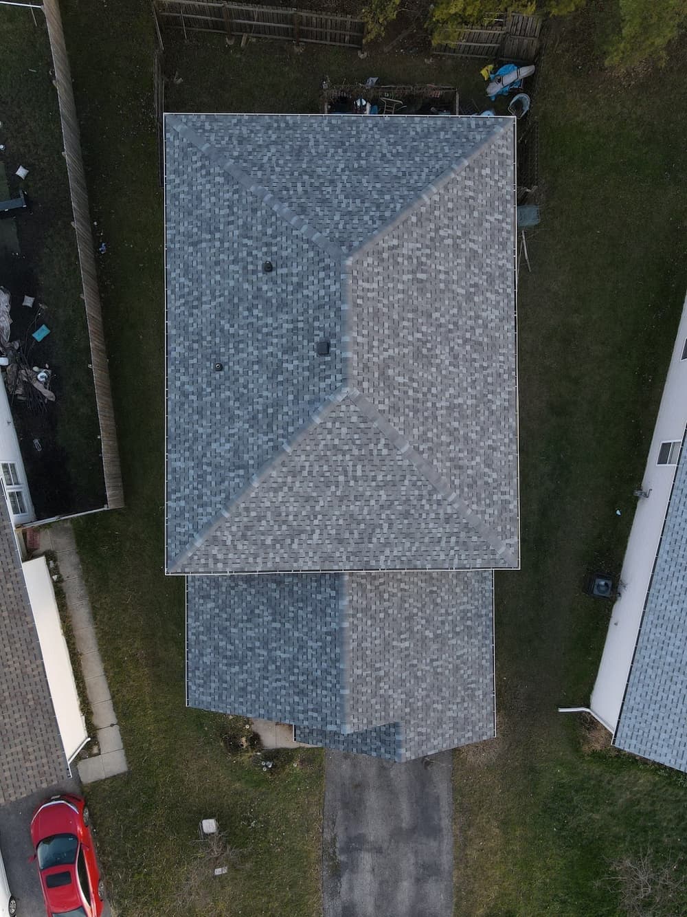 Aerial view of a residential roof with gray shingles surrounded by green grass and driveways.