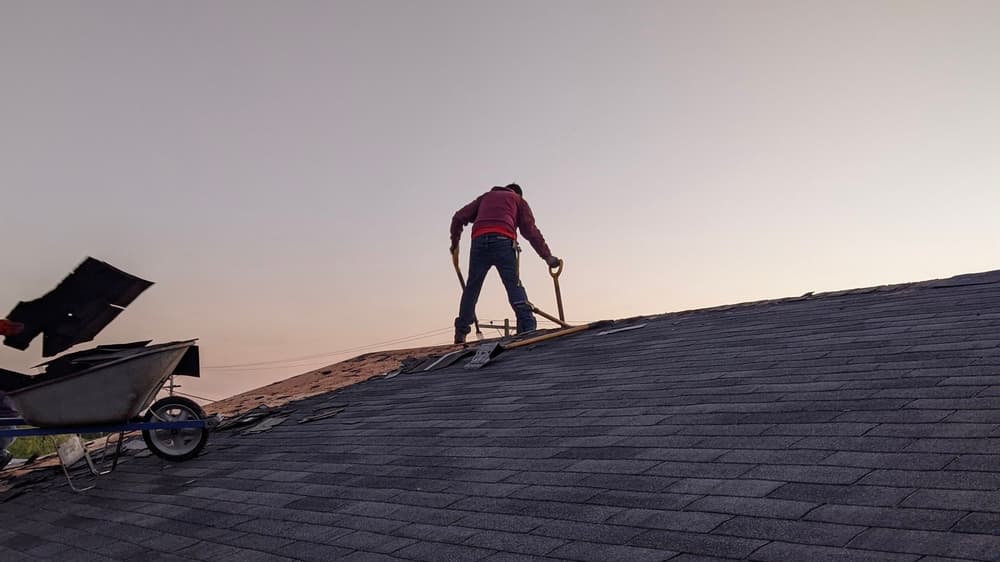 Person repairing a roof at sunset, with tools and a wheelbarrow nearby.