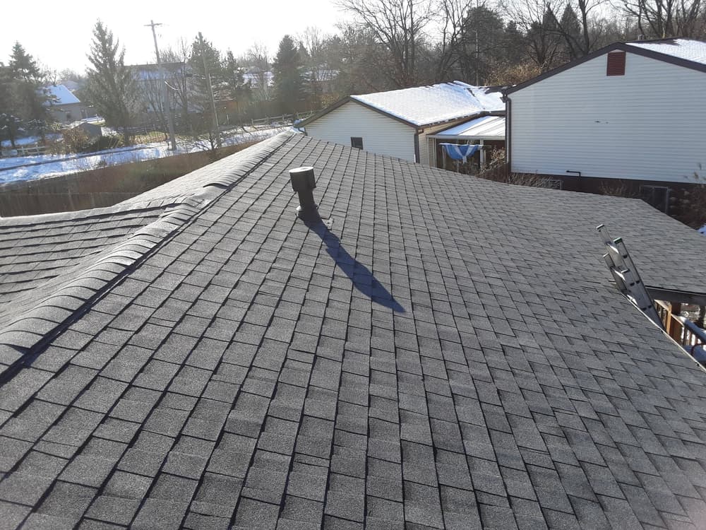 Aerial view of a black shingle roof with a chimney against a snowy suburban background.
