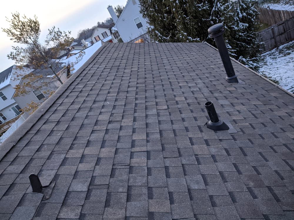Overview of a snow-dusted asphalt shingle roof with vent pipes near residential houses.