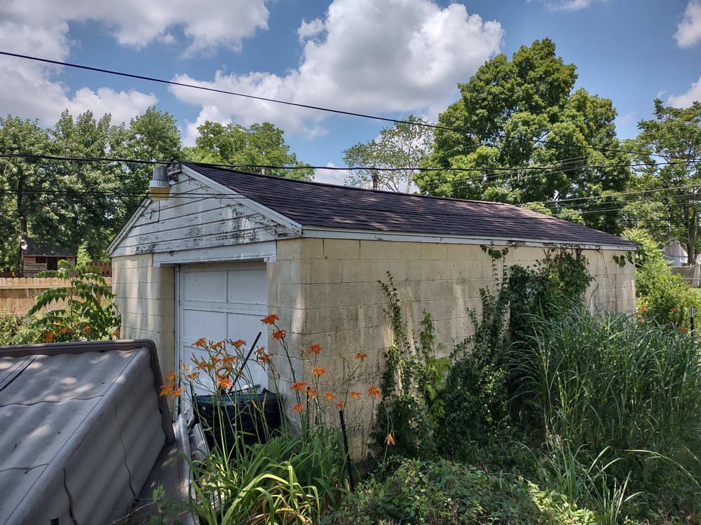 Abandoned garage with overgrown plants and trees under a blue sky with clouds.