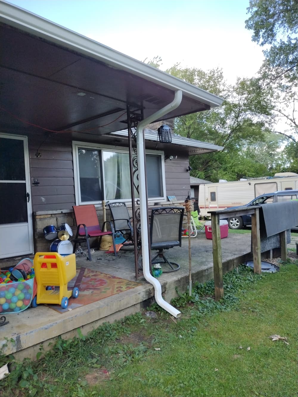 White downspout draining water from roof near a porch with toys and chairs. Outdoor setting.