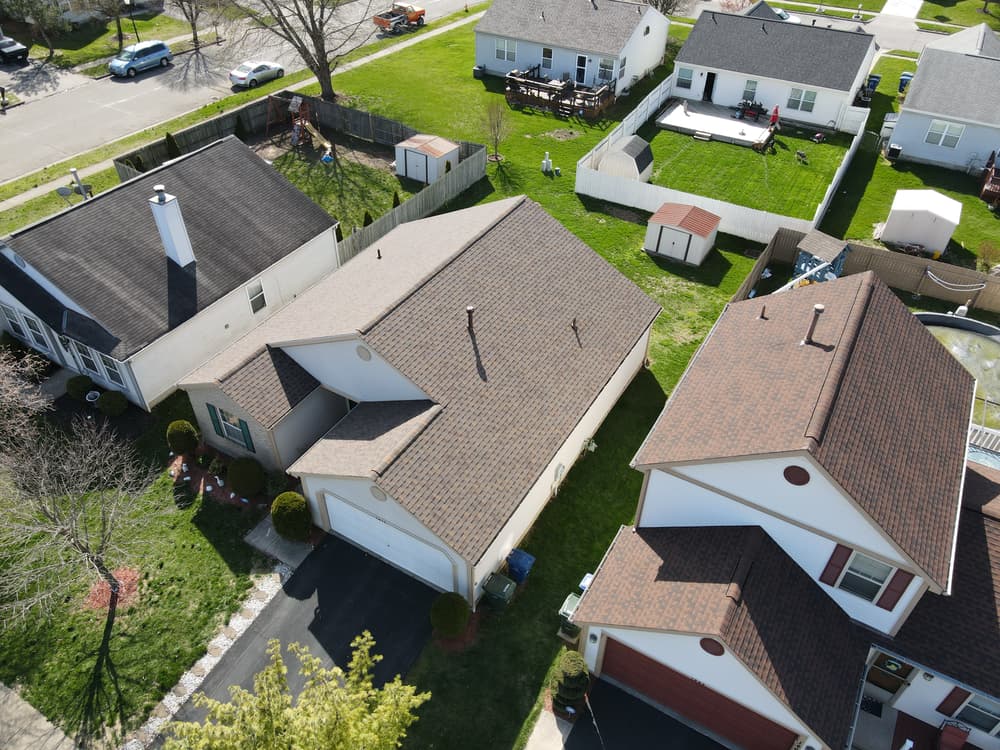 Aerial view of suburban homes with landscaped yards and asphalt roofs. Clear blue sky above.